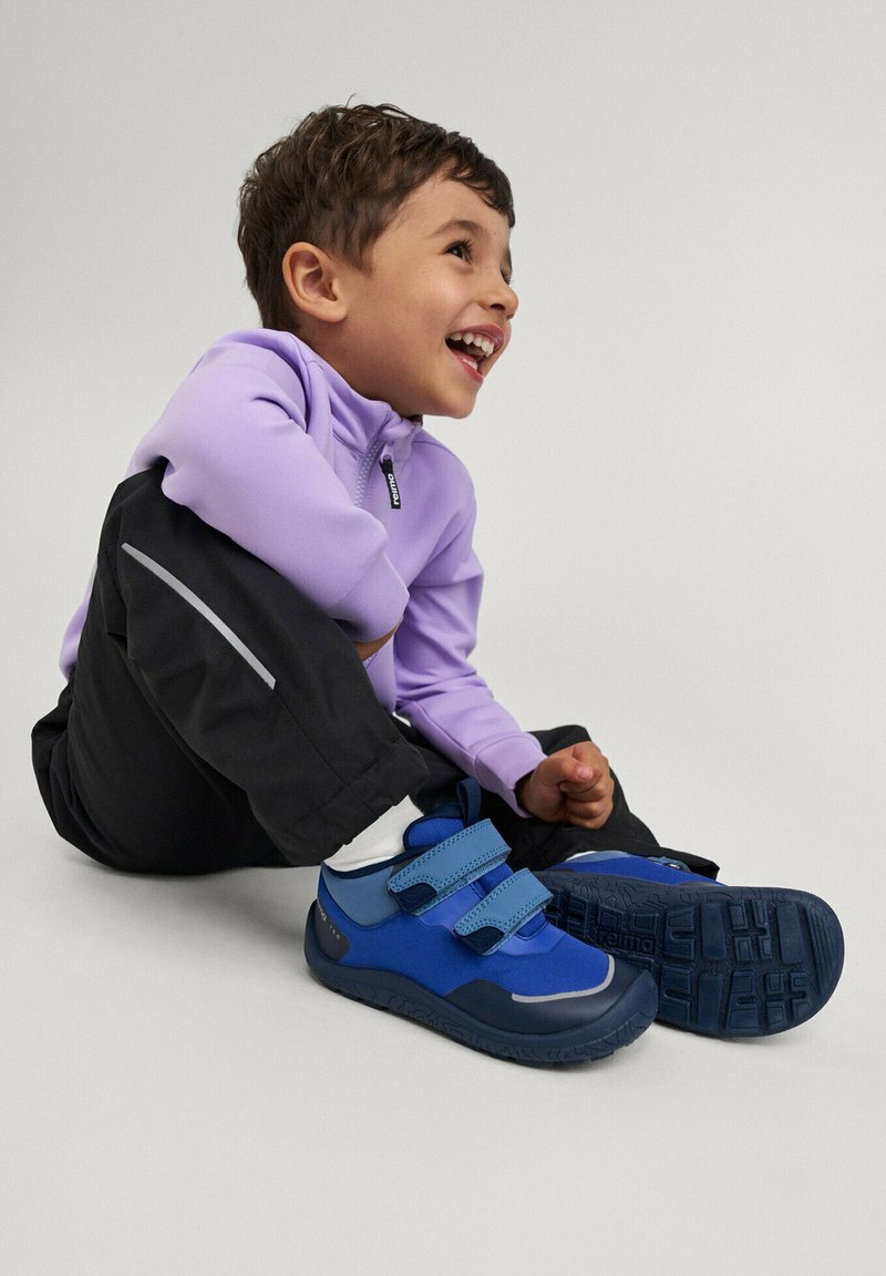 Młody chłopiec śmieje się, siedząc na podłodze, ubrany w jasnofioletową kurtkę, czarne spodnie i niebieskie trampki na rzepy.