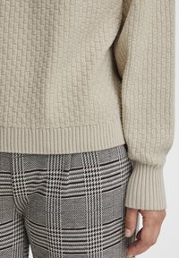 Pull en tricot texturé beige avec des poignets côtelés, associé à un pantalon noir et blanc à motifs comportant des carreaux et un design pied-de-poule.