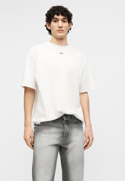 Νέος άνδρας με κατσαρά μαλλιά που φοράει λευκό υπερμεγέθη μπλουζάκι με το κείμενο "OFF" και ξεθωριασμένο γκρι τζιν, όρθιος μπροστά σε απλό φόντο.