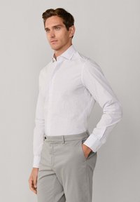 Camicia bianca a maniche lunghe con motivo a quadretti grigio chiaro e rosso, abbinata a pantaloni grigi con tasca frontale.