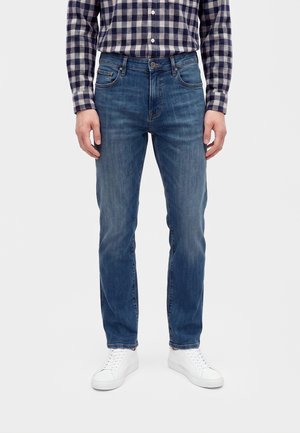 Mann trägt blaue gerade geschnittene Jeans, weiße Sneakers und ein blau-weiß kariertes Hemd mit Knöpfen, steht vor einem einfarbigen Hintergrund.