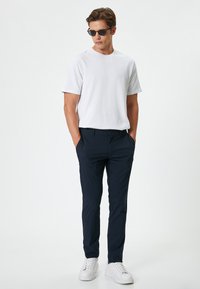 Camiseta blanca de cuello redondo, pantalones ajustados navy y zapatillas blancas. El modelo lleva gafas de sol y tiene las manos en los bolsillos, de pie frente a un fondo liso.