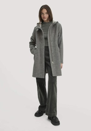 Grauer Wollmantel mit Kapuze, ausgestattet mit einem Reißverschluss und seitlichen Taschen. Kombiniert mit einem grauen Oberteil und dunkelgrünen Cordhosen. Schwarze Schuhe.