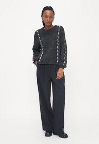 Pulover gros negru cu cusături decorative albe, guler rotund și umeri lăsați. Asortat cu pantaloni negri largi și pantofi negri.