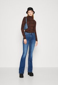 Rjava puma z vzorcem turtleneck majica, modri široki jeans, črne platform čevlje in črna vedro kapa. Model drži majhno modro torbico.