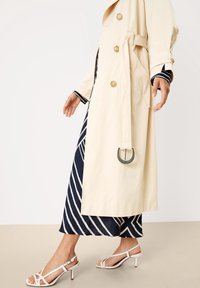 Gabardina beige con botones dorados, cinturón de lazo y un adorno metálico. Lleva puesta sobre un vestido de rayas azul marino y combinado con sandalias blancas de tiras.