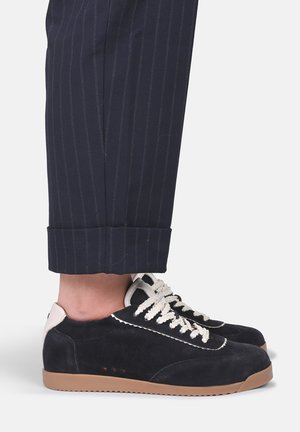 Sneakers in pelle scamosciata nera con lacci bianchi e suole in gomma color cuoio, indossate con pantaloni blu navy a righe sottili risvoltati sopra la caviglia.