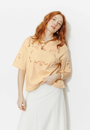 Femme aux cheveux roux portant un haut pêche à découpes avec le texte « JOY », une jupe blanche, des colliers superposés, et posant devant un fond blanc.