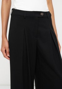 Personne portant un pantalon noir plissé taille haute avec une ceinture boutonnée et un haut blanc, montré du milieu du torse aux cuisses.