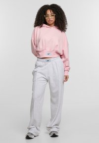 Cropped hoodie rosa chiaro con una targhetta logo frontale, abbinato a pantaloni della tuta grigi a vita alta e scarpe da ginnastica nere e grigie.