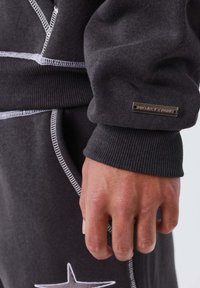 Sudadera negra con puños acanalados, que presenta costuras en contraste y una etiqueta metálica con logotipo. La mano agarra el puño con un acabado texturizado.