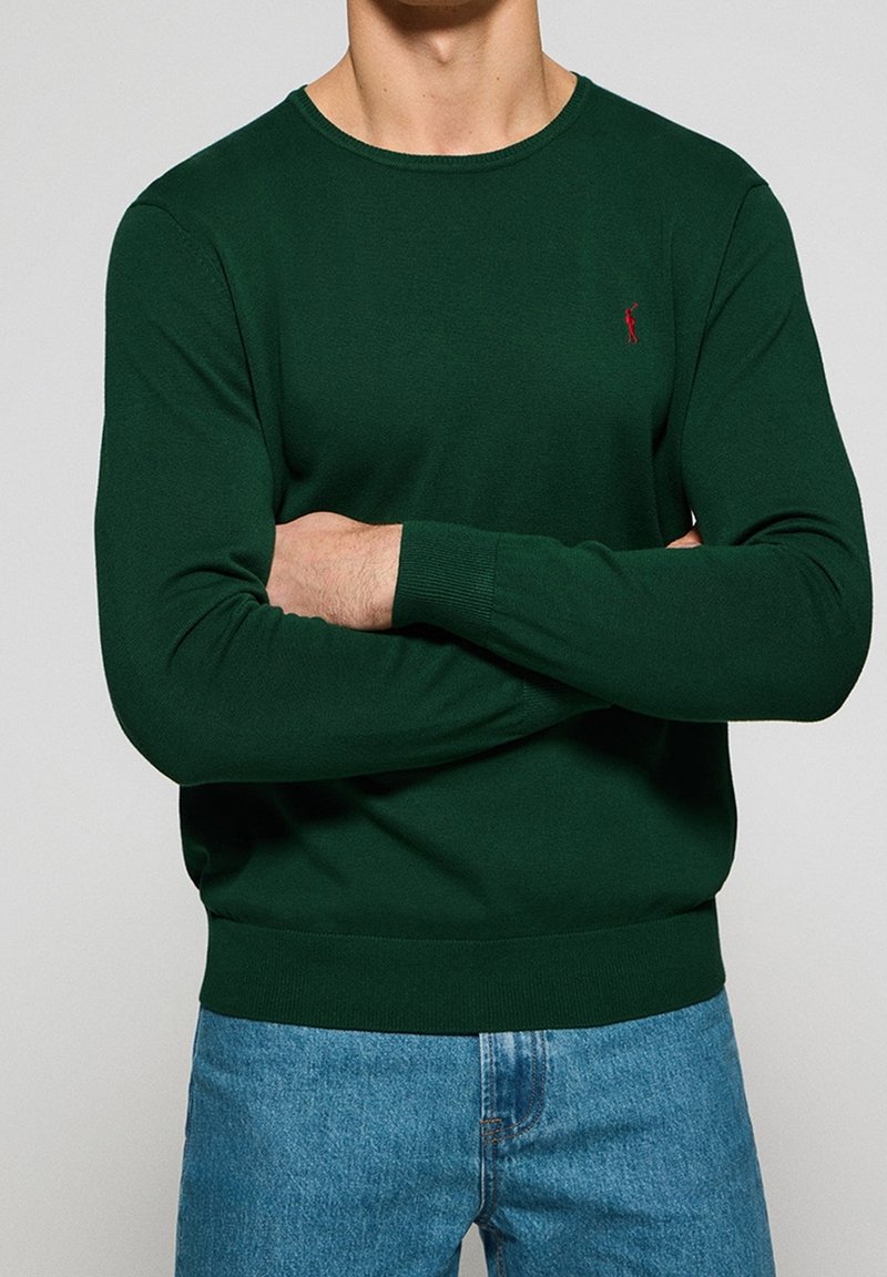 Ciemnozielony sweter dzianinowy z okrągłym dekoltem, ściągaczami przy mankietach i u dołu. Zawiera małe czerwone logo na klatce piersiowej. Połączony z niebieskimi dżinsami.