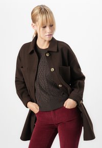 Donna con capelli biondi che indossa un cappotto buttoned marrone scuro, un maglione a maglia a trecce e pantaloni di velluto a coste rossi, con le mani in tasca, che guarda in basso.