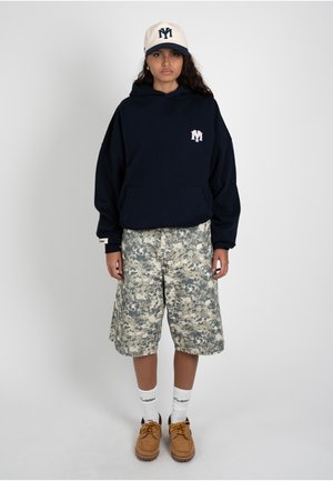 Jeune personne portant une casquette blanche, un sweat à capuche noir, un short camouflage, des chaussettes blanches et des chaussures marron, debout sur un fond uni.