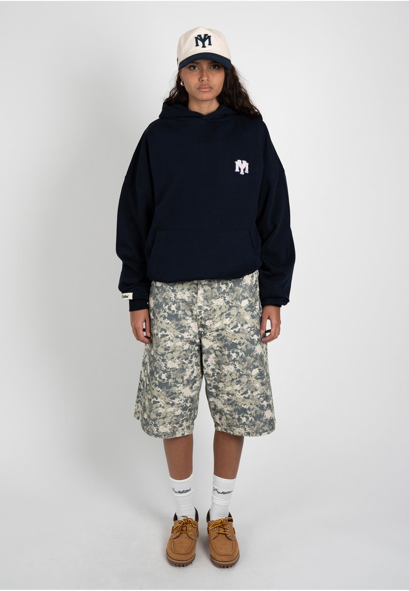 Jeune personne portant une casquette blanche, un sweat à capuche noir, un short camouflage, des chaussettes blanches et des chaussures marron, debout sur un fond uni.