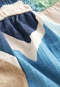 Tissu multicolore présentant des motifs géométriques dans des tons de bleu, de marine, de crème et de vert clair. Texturé, léger, avec une taille froncée.