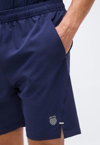 Κοντινό πλάνο ατόμου που φοράει ναυτικό μπλε αθλητικό σορτς με ελαστική μέση, το χέρι στην πλαϊνή τσέπη, λογότυπο K-Swiss κοντά στο τελείωμα.