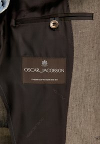 Braunes Stofffutter einer Jacke mit einem Logoetikett "OSCAR JACOBSON" und "SCHWEDISCHES HANDWERK SEIT 1903"; verfügt über einen Knopf und eine strukturierte Oberfläche.