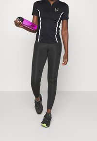 Svart sporttröja med vita detaljer, matchad med svarta leggings. Håller en transparent lila vattenflaska. Svarta sportskor med gröna detaljer.