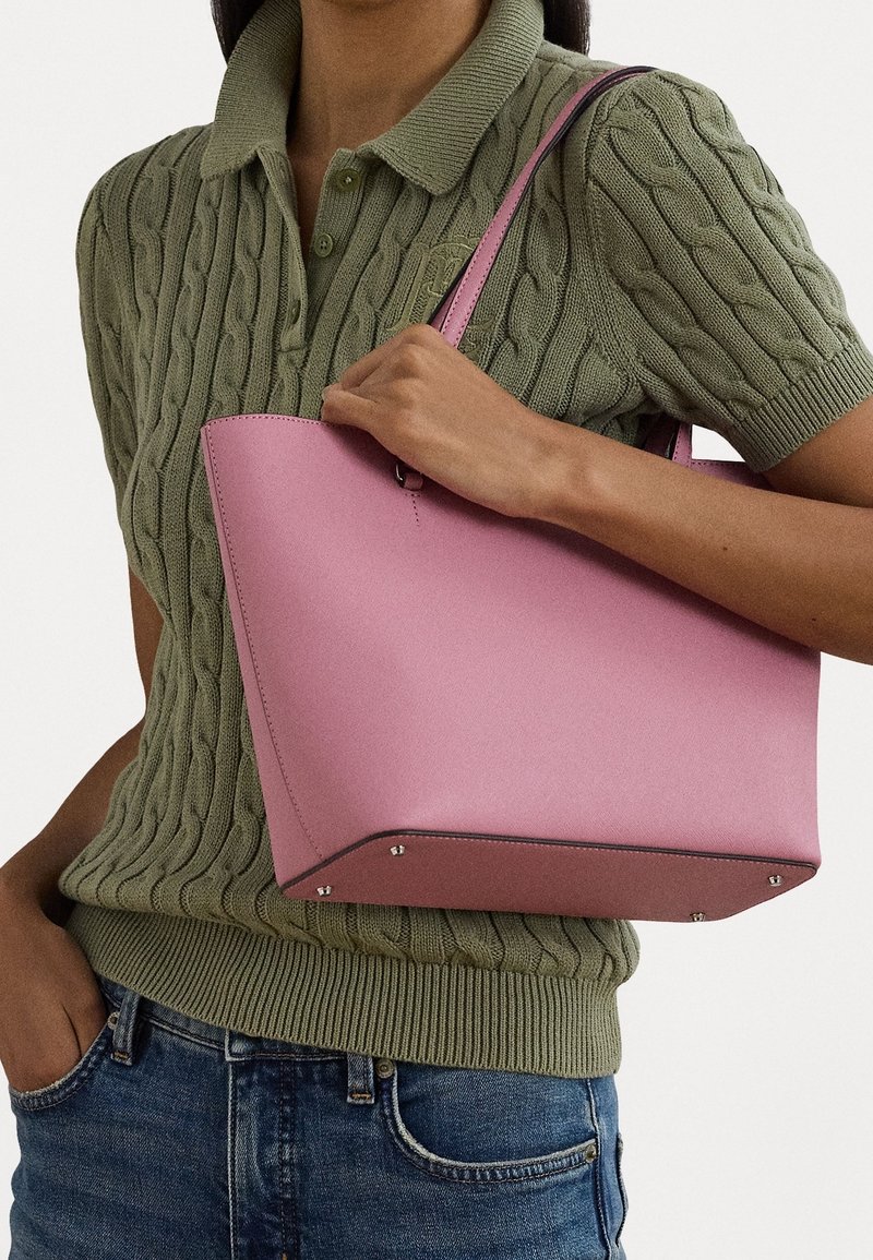 Žena v olivově zeleném pleteném polo tričku s krátkým rukávem, držící strukturovanou růžovou koženou kabelku se stříbrnými hroty, v modrých džínách.
