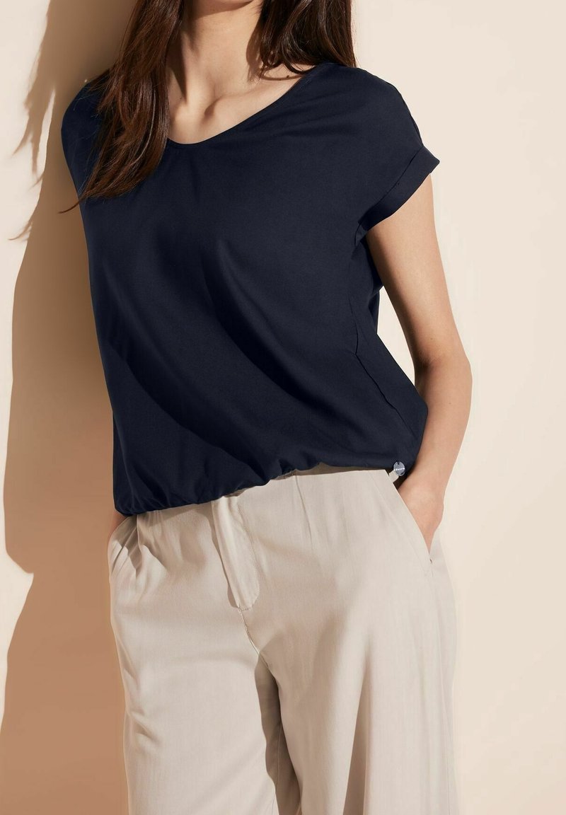 Femme portant un chemisier bleu marine à manches courtes, rentré dans un pantalon large à taille haute beige clair, debout les mains dans les poches.