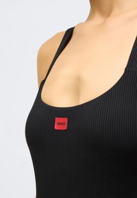 Fekete bordázott trikó, bal oldalon egy piros, téglalap alakú HUGO feliratos címkével. Vékony pántokkal és kerek nyakkivágással.