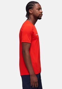 Rød idræts t-shirt med korte ærmer, glat tekstur og subtilt mesh-design. Har et logo på brystområdet, iført med navy shorts.