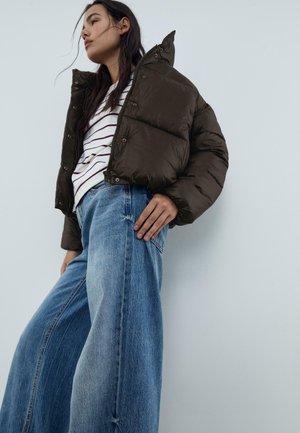 Frau in einer cropped, dunkelbraunen Pufferjacke, gestreiftem weißen Shirt und weit geschnittenen blauen Jeans, die vor einer schlichten hellgrauen Wand posiert.