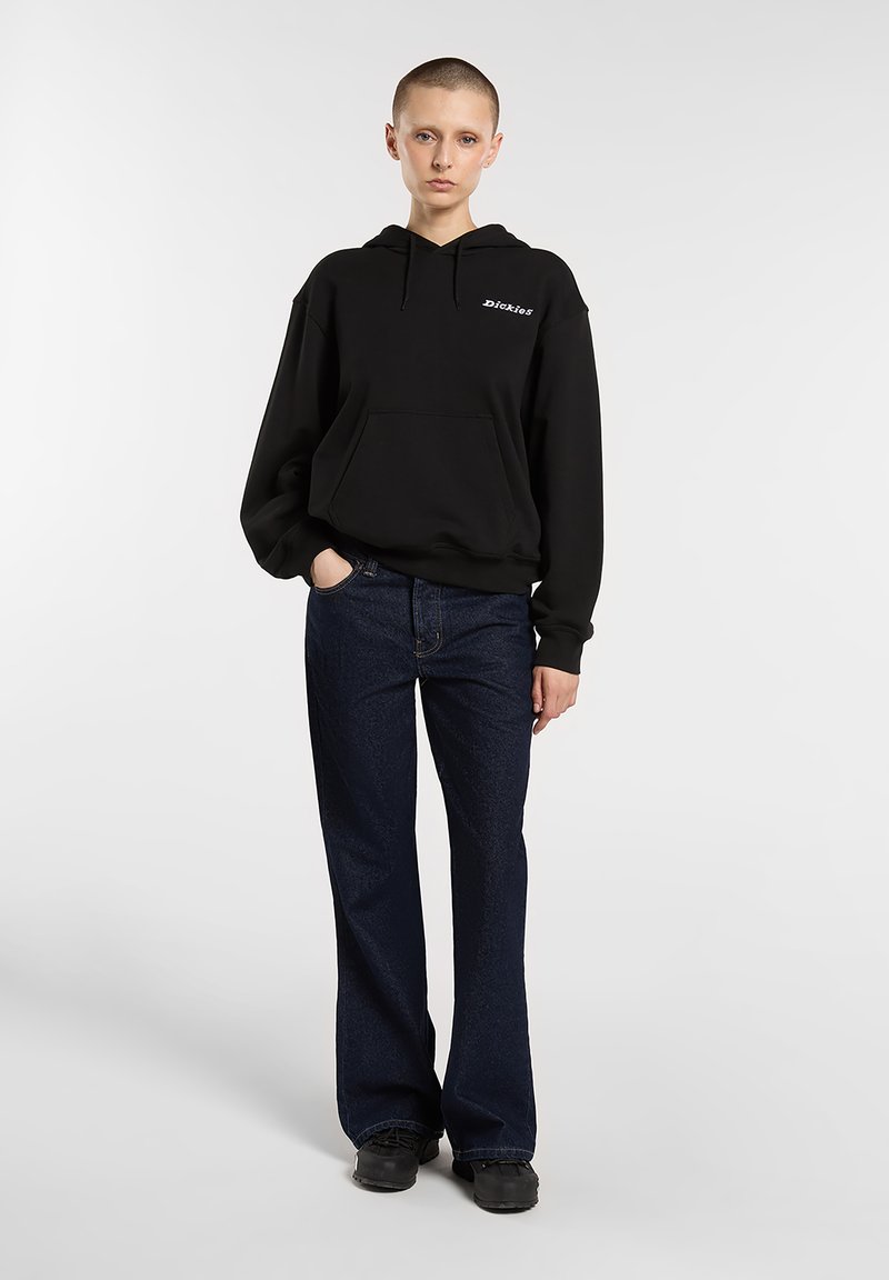 Zwarte capuchontrui met een voorzak en geborduurd logo, gecombineerd met uitlopende donkere spijkerbroeken en zwarte schoenen. Over het geheel een gladde textuur.