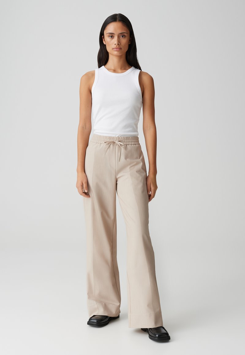 Pantalons larges beiges avec taille élastique et cordon de serrage, associés à un débardeur blanc ajusté. Texture lisse et design minimal.