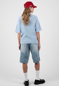 Femme portant une casquette rouge, une chemise à manches courtes à motifs bleu clair, un long short en jean, des chaussettes blanches et des chaussures noires, debout de dos.