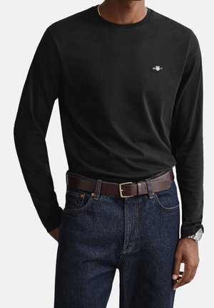 Mężczyzna ubrany w czarną koszulę z długim rękawem z małym logo na piersi, ciemnoniebieskie dżinsy, brązowy skórzany pasek i srebrny zegarek na nadgarstku.