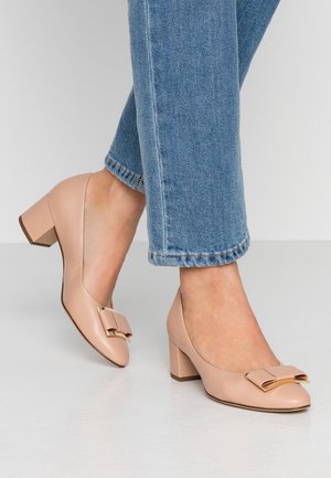 Beżowe skórzane buty na obcasie blokowym z zaokrąglonym noskiem i ozdobną kokardką z przodu. Noszone z skróconymi niebieskimi dżinsami.