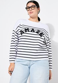 Maglione a righe bianche e nere con la parola "AMAZE" in nero, caratterizzato da una vestibilità morbida e maniche lunghe, abbinato a jeans azzurri chiari.