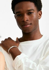 Zilveren armband om een pols met een donkere huid, gecombineerd met een wit overhemd met lange mouwen. De achtergrond is effen wit voor contrast.