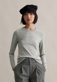 Langärmliges graues Oberteil mit rundem Halsausschnitt, kombiniert mit grauen gestreiften Hosen. Eine schwarze Baskenmütze und eine silberne Lagenkette runden das Outfit ab.