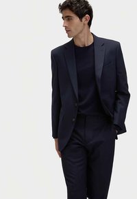 Navy blue suit with a slim fit, featuring a notched lapel and single button closure. Soft fabric, smooth texture, worn over a matching top.