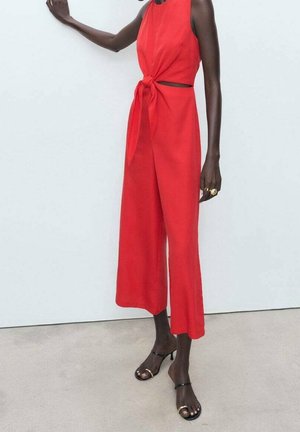Combinaison rouge sans manches, avec un détail noué à la taille, jambes larges et découpes sur les côtés. Associée à des talons noirs à lanières.