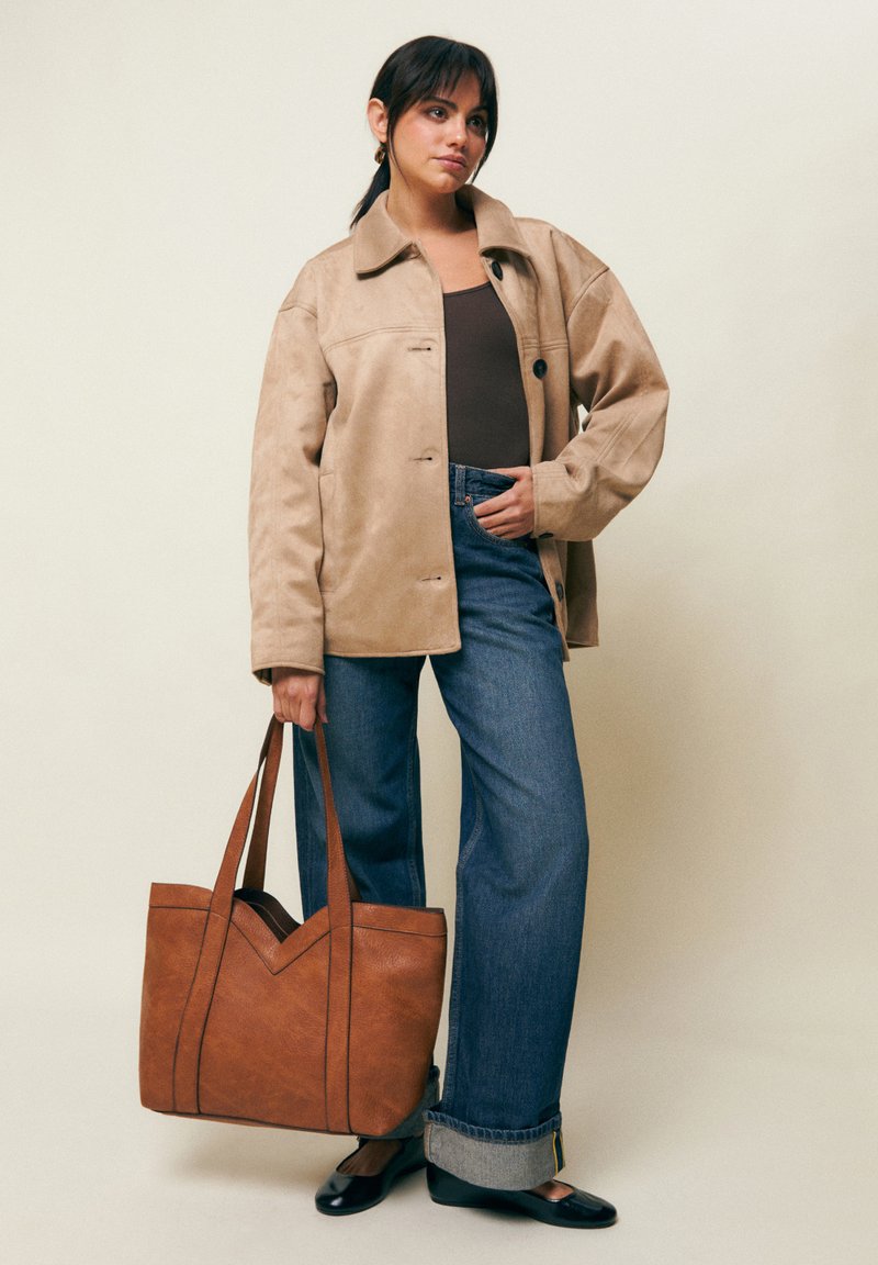 Mujer con chaqueta beige oversize, top oscuro, vaqueros azules remangados, bailarinas negras, sosteniendo un gran bolso tote de cuero marrón sobre un fondo liso.