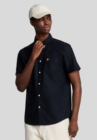 Camicetta blu navy a maniche corte con taschino sul lato sinistro e dettagli logo giallo. Tessuto di cotone, texture liscia e bottoni bianchi.
