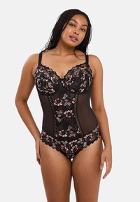 Femme avec des cheveux tressés portant un body en dentelle florale noire avec des panneaux en maille, posant devant un fond clair et uni.