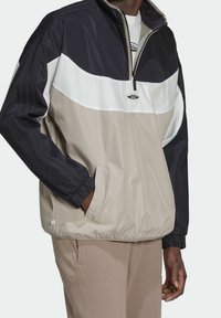 Man som bär en färgblockerad vindtålig jacka i svart, vitt och beige med halvdragkedja och handen i fickan, ihop med beige byxor.