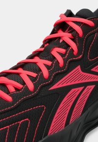 Μαύρο αθλητικό παπούτσι με φωτεινές κόκκινες κορδόνες και γεωμετρικά λογότυπα της μάρκας σε κόκκινο χρώμα, κοντινή λήψη λεπτομερειών του επάνω μέρους και της ραφής.