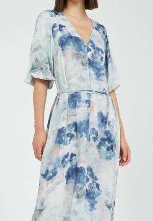 Frau trägt ein Kleid mit Gürtel mit blauem, grauem und pfirsichfarbenem Aquarell-Blumenmuster und gepufften kurzen Ärmeln, vor einfachem Hintergrund.