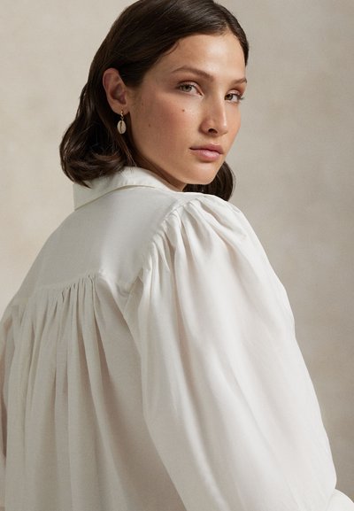 Hvit, overdimensjonert bluse med foldedetaljer på baksiden, med en strukturert krage og mykt, lett stoff.