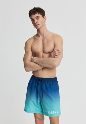 Uomo senza maglietta con capelli corti castani, in piedi con le braccia incrociate, indossa pantaloncini da bagno con sfumatura dal blu al turchese con scritto "TERRANOVA".