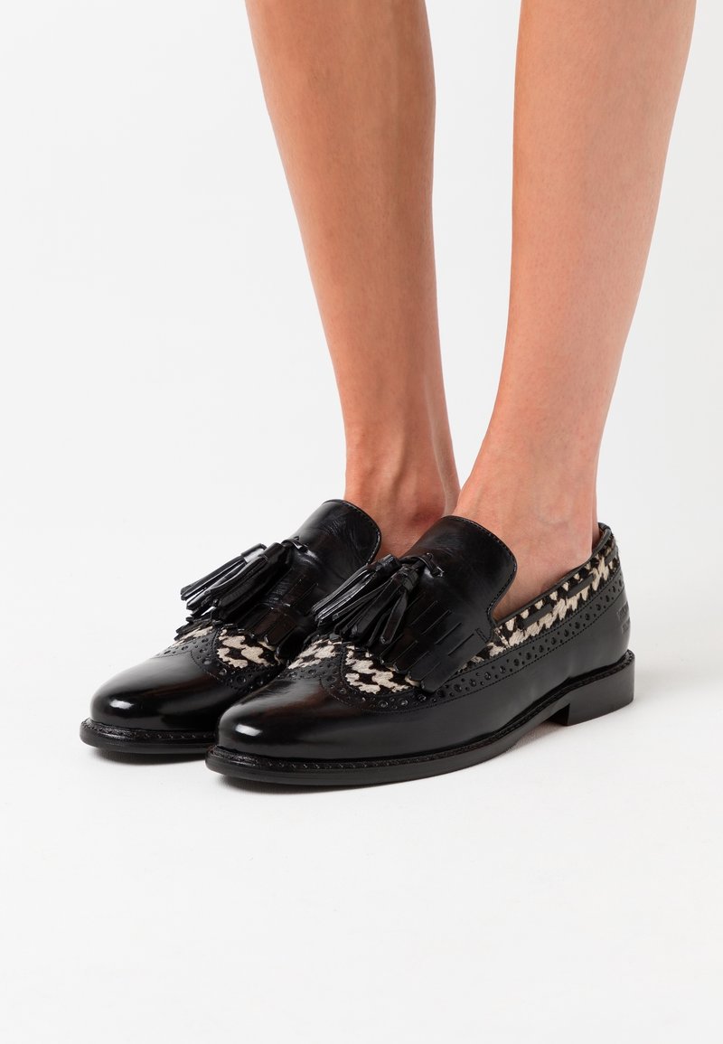 Mocassins noirs en cuir à pampilles avec des inserts en tissu à motifs portés pieds nus sur fond blanc.