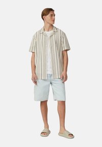 Korte mouw crèmekleurig shirt met verticale strepen, witte t-shirt eronder, lichtblauwe denim shorts en beige schuifsandalen.