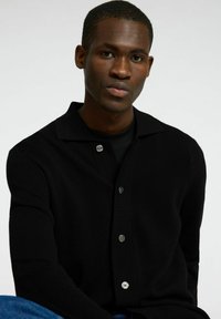 Svart cardigan med krage, med knappelukking, ribbestruktur og en tettsittende design. Båret over en svart topp.