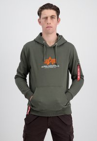 Felpa verde oliva con tasca frontale, cappuccio con cordino e logo arancione. Presenta un'etichetta rossa sulla manica, polsini e orlo a costine.